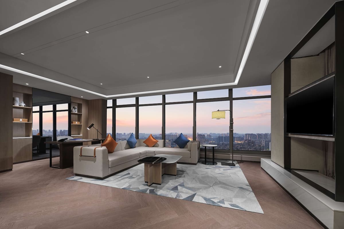 A modern living room with a sofa, coffee table, and TV, large windows with a city view, and built-in shelving.