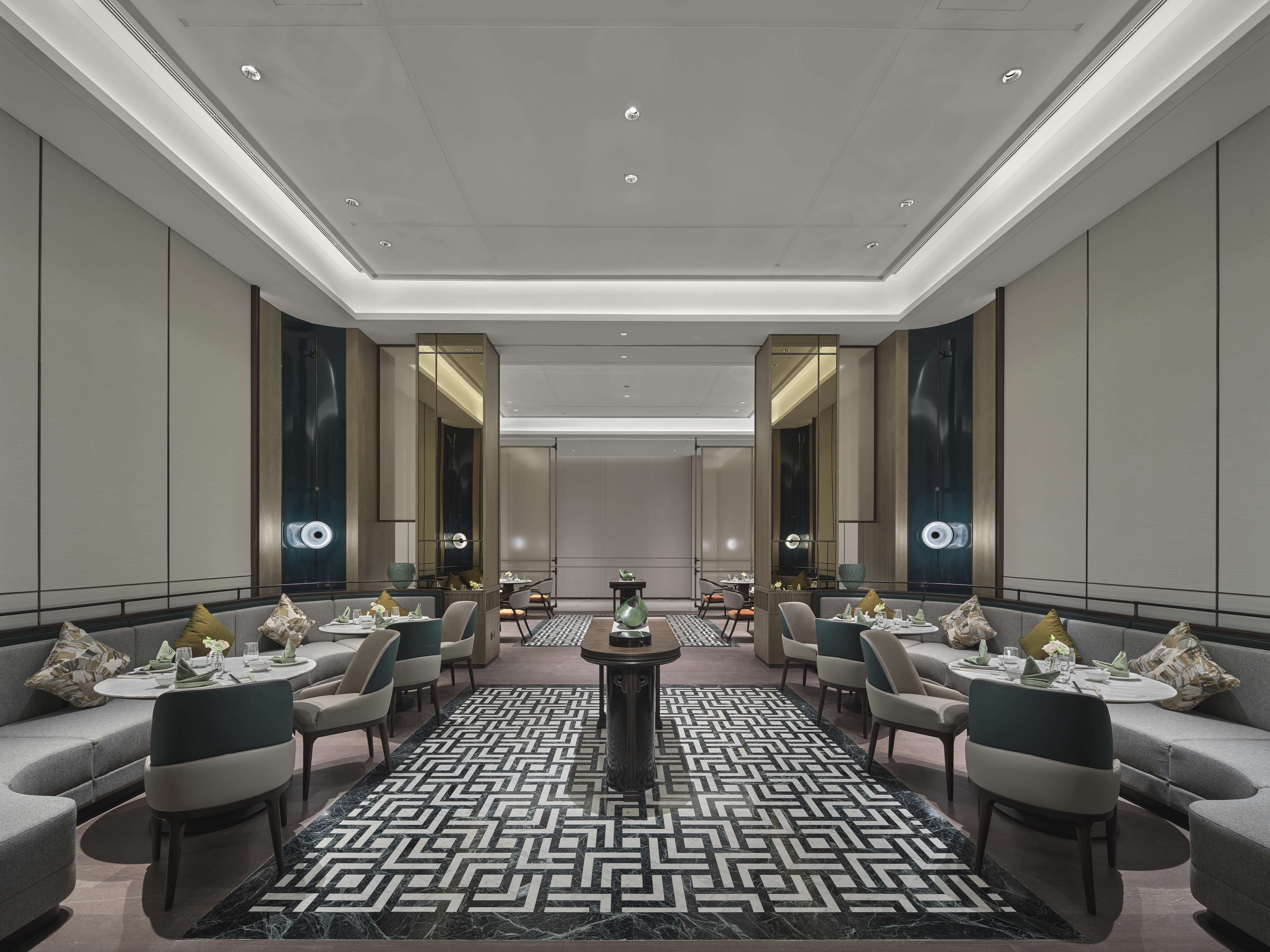 A modern hotel lobby with a symmetrical layout, featuring a central round table, seating arrangements, and a patterned carpet.