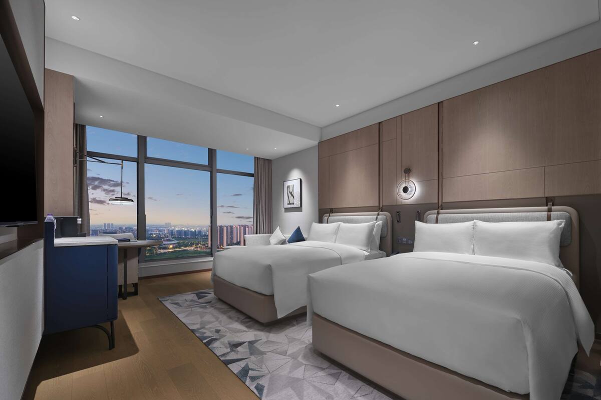 A modern hotel room with two beds, a desk, and a large window offering a city view.