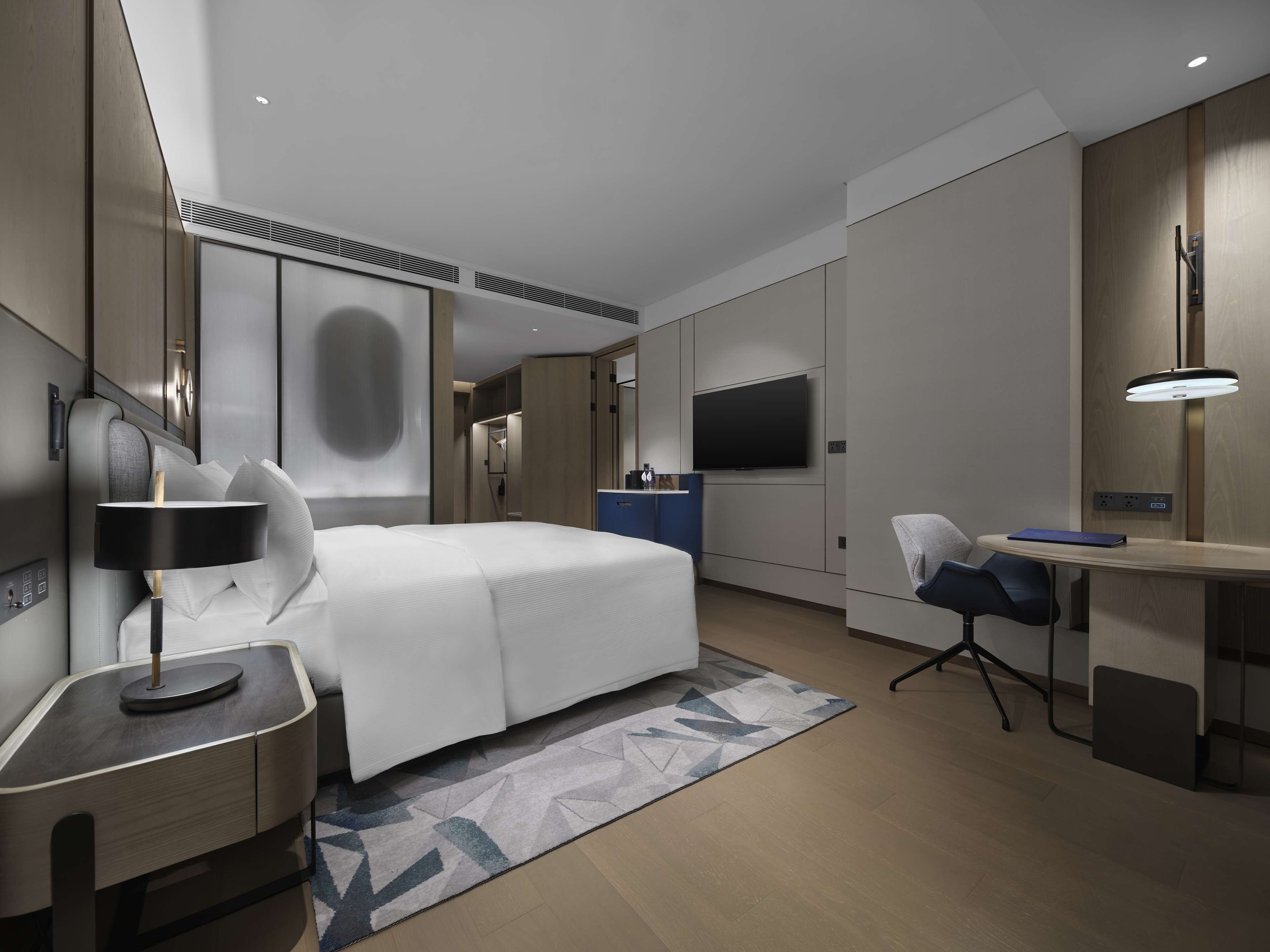 A modern hotel room with a bed, desk, chair, TV, and a lamp.