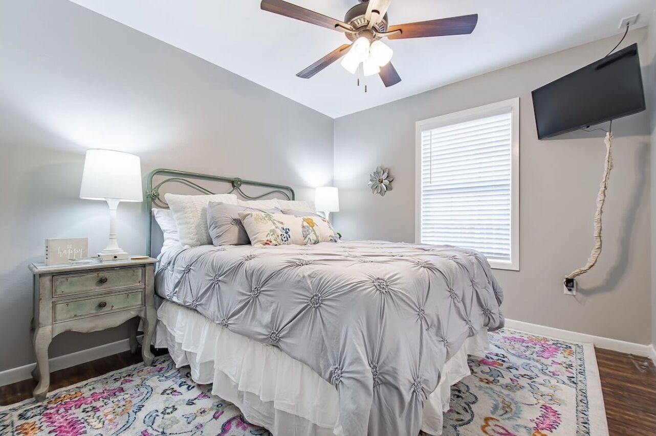 A bedroom with a bed, nightstand, lamp, ceiling fan, TV, and window with blinds.