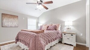 Un dormitorio con cama, ventilador de techo, mesita de noche con lámpara y ventana con persianas.