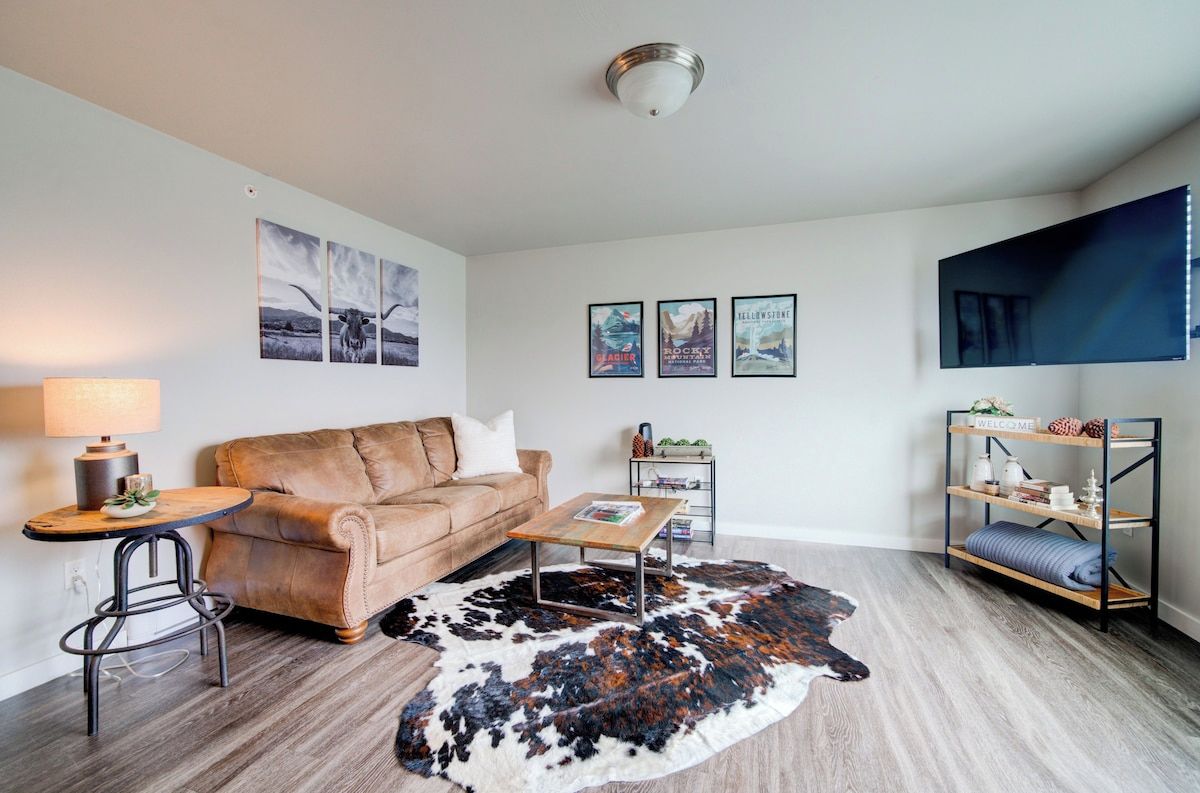A living room with a brown leather sofa, a round coffee table, a cowhide rug, a TV mounted on the wall, and a small side table with a lamp.