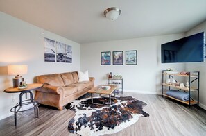 A living room with a brown leather sofa, a round coffee table, a cowhide rug, a TV mounted on the wall, and a small side table with a lamp. - King Bed Condo | Country Views | 5 Min From MSU (Bozeman)