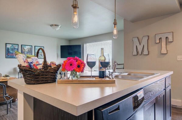 A kitchen with a central island, a sink, and a dishwasher. There are hanging light fixtures, a wall-mounted TV, and a basket with groceries and flowers. - King Bed Condo | Country Views | 5 Min From MSU (Bozeman)