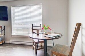 A dining area with a round table, chairs, a vase with flowers, and a window with blinds. - King Bed Condo | Country Views | 5 Min From MSU (Bozeman)