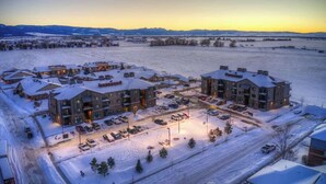 Un quartier résidentiel enneigé, avec des immeubles d’appartements modernes, un parking et une vue dégagée sur les montagnes lointaines.