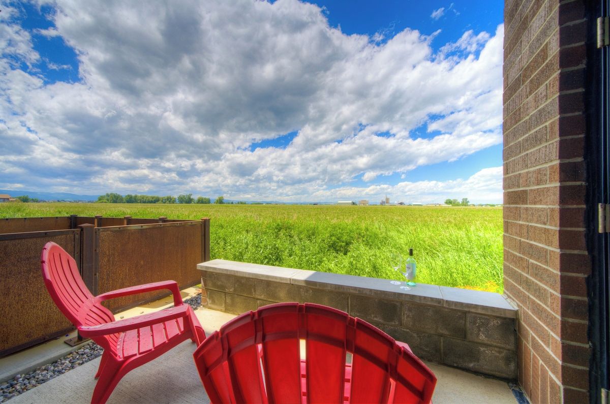 Un balcon avec des chaises rouges, une bordure en béton et une vue sur un champ verdoyant sous un ciel partiellement nuageux.