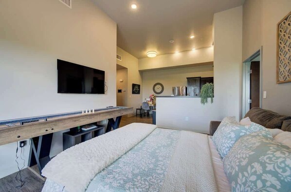 A modern bedroom with a large bed, a desk with a flat-screen TV, and a kitchenette in the background.