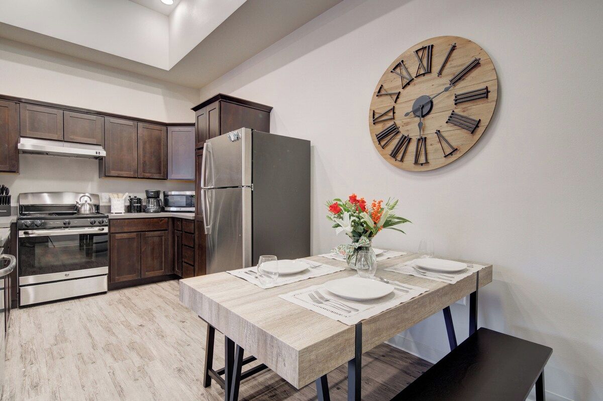 A modern kitchen with dark wood cabinets, stainless steel appliances, and a wooden dining table set for two with a vase of flowers.