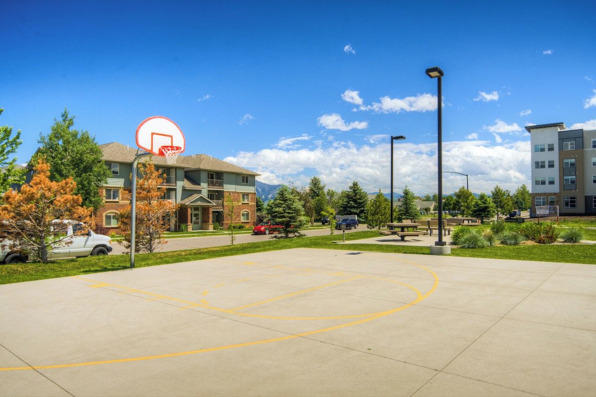 Una cancha de baloncesto al aire libre con aro, rodeada de edificios, árboles y cielo despejado.