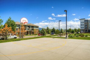 An outdoor basketball court with a hoop, surrounded by buildings, trees, and a clear sky.