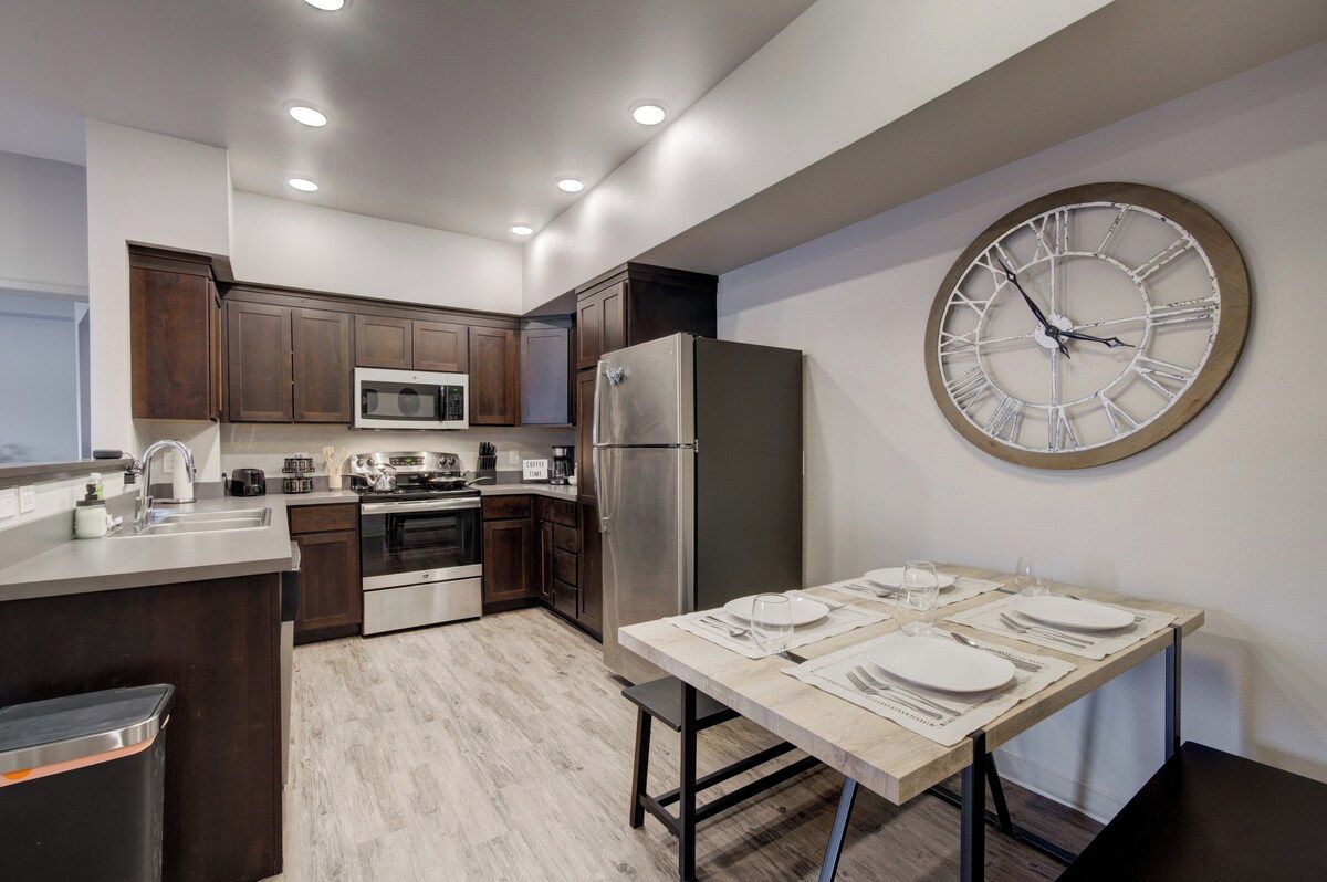 A modern kitchen with dark wooden cabinets, stainless steel appliances, a large wall clock, and a dining area set for four.