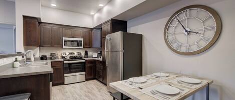 A modern kitchen with dark wooden cabinets, stainless steel appliances, a large wall clock, and a dining area set for four.