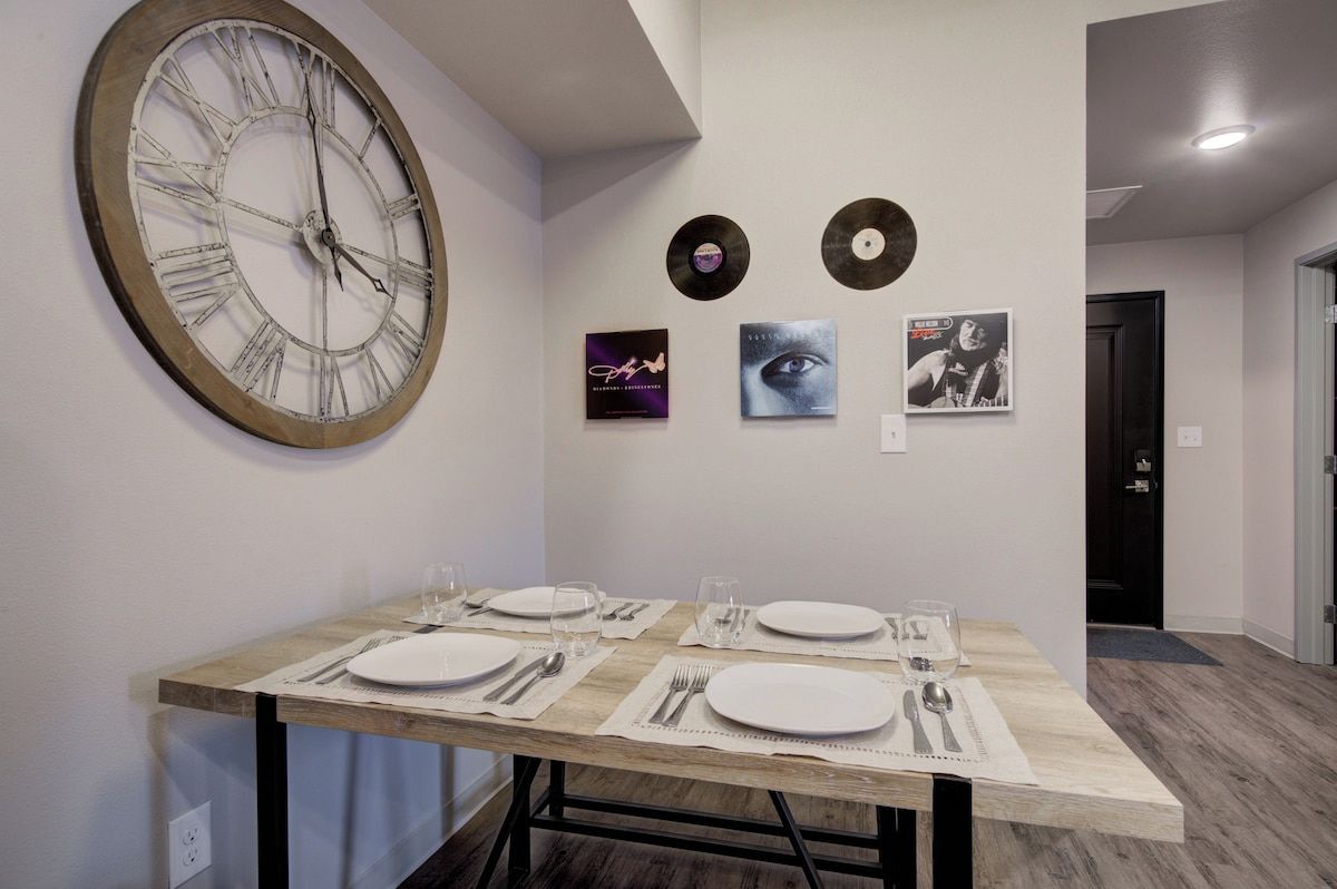 A dining area with a wooden table set for four, a large wall clock, and three framed items on the wall.