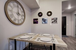 A dining area with a wooden table set for four, a large wall clock, and three framed items on the wall. - King Bed | Country Music in the Big Sky (Bozeman)