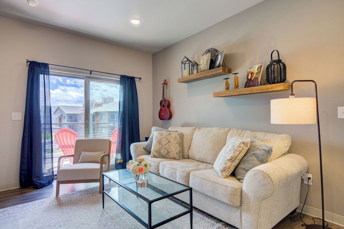 A living room with a sofa, a glass coffee table, a chair, a guitar, and shelves with decorative items.