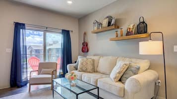 A living room with a sofa, a glass coffee table, a chair, a guitar, and shelves with decorative items.