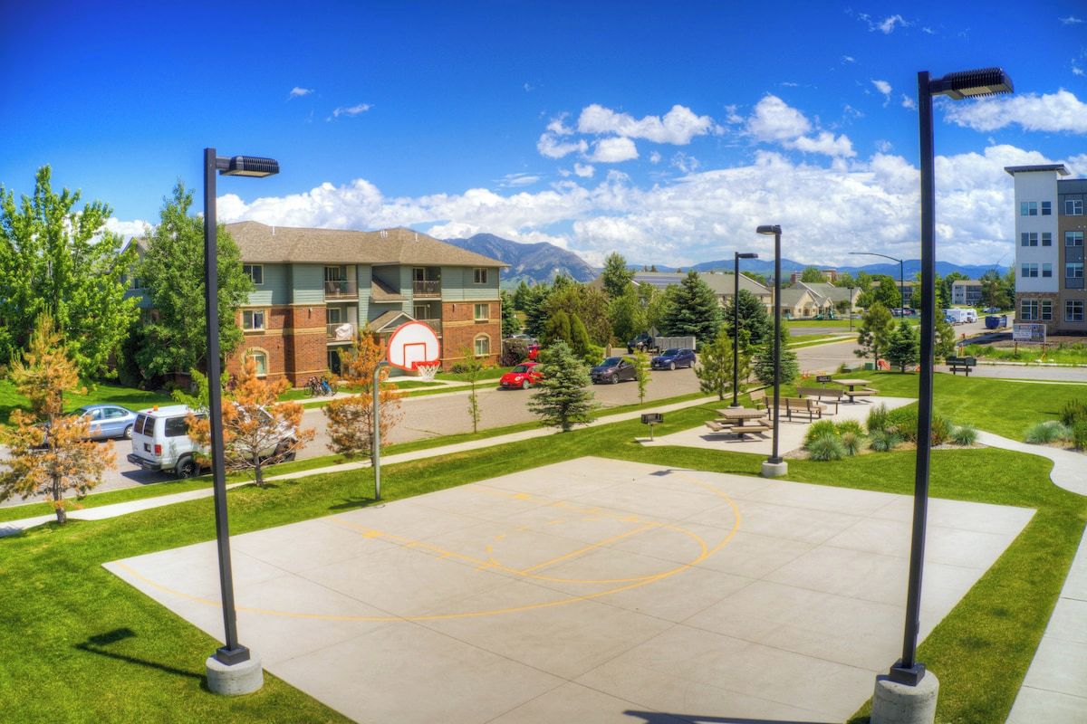 A basketball court with a building in the background, surrounded by trees and parked cars.