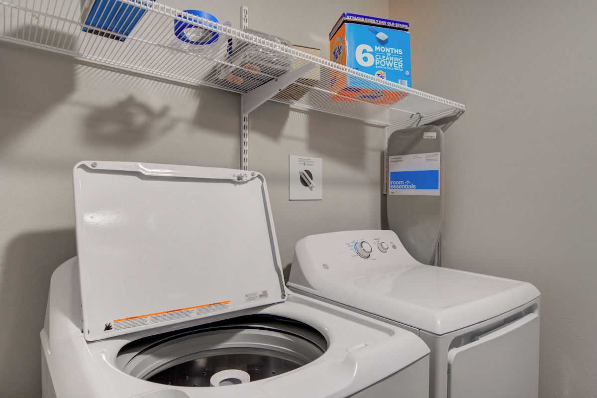A laundry room with a front-loading washing machine, a shelf with cleaning supplies, and a sign indicating the room's use.