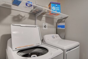 Une buanderie équipée d’un lave-linge à chargement frontal, d’une étagère contenant des produits de nettoyage et d’un panneau indiquant l’usage de la pièce.
