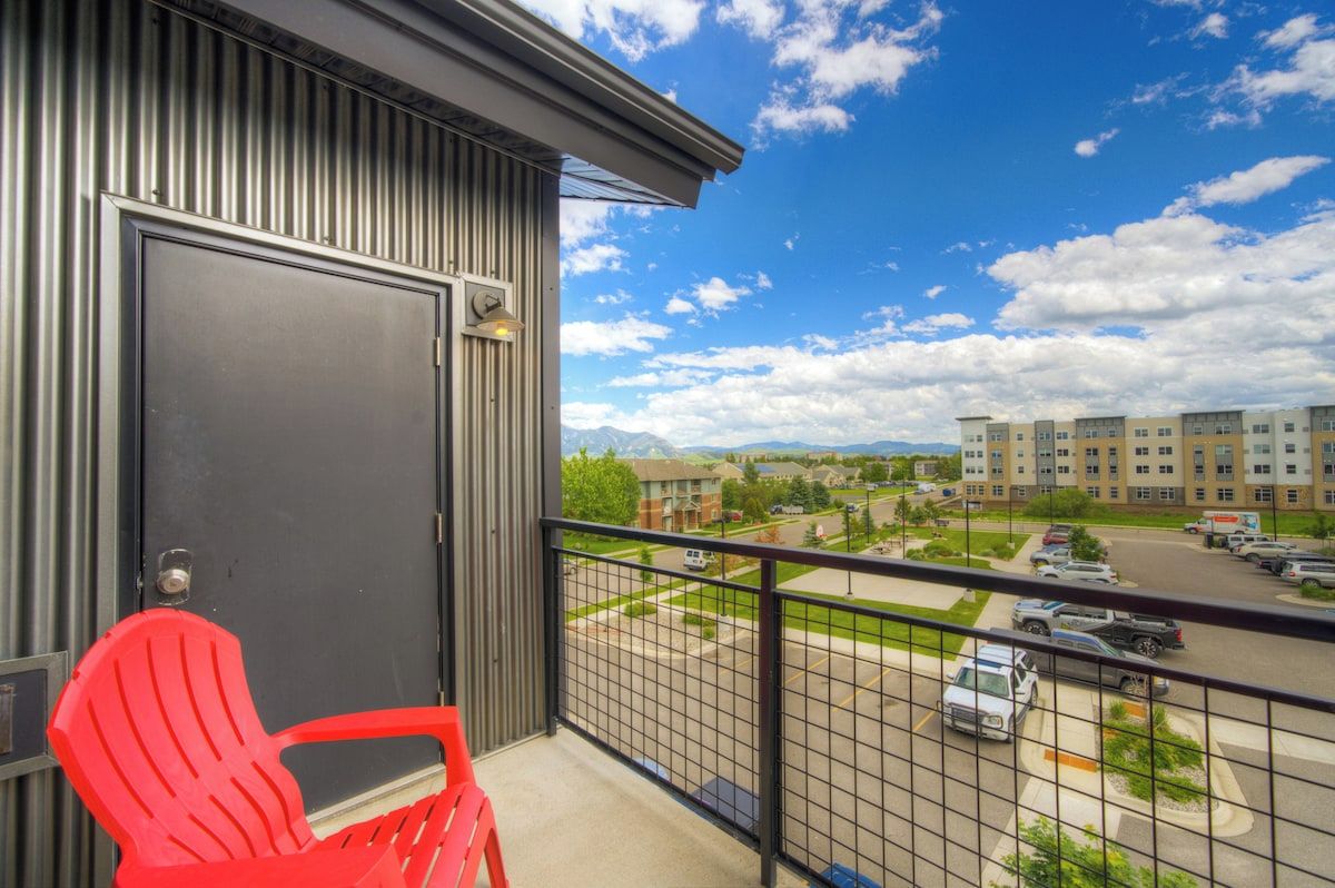 A balcony with a red chair, a metal door, and a view of a parking lot and buildings.
