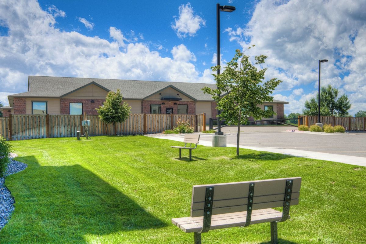 A building with a paved driveway, a wooden fence, a bench, and a tree.