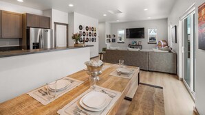 Una cocina moderna con refrigerador integrado, mesa de comedor de madera preparada para cuatro comensales y una sala de estar con sofá y sistema de entretenimiento.