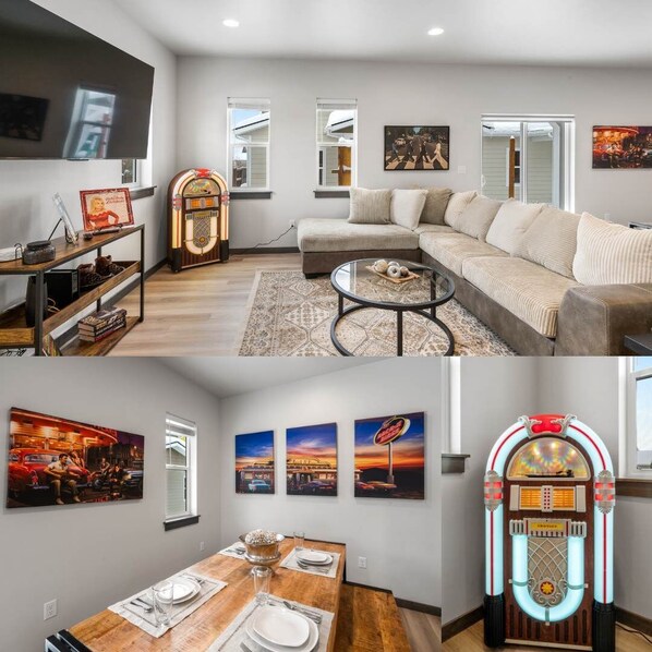 A living room with a sofa, coffee table, and jukebox. There is a dining area with a table set for two and artwork on the walls. - Heart of Bozeman | Juke Box Jet Away (Bozeman)