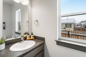 A bathroom with a white sink, dark countertop, and a large mirror. There are toiletries on the counter and a window with a view of a house and a fence. - Heart of Bozeman | Juke Box Jet Away (Bozeman)