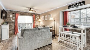 Un soggiorno moderno con un divano grigio, un tavolino da caffè bianco e un'area pranzo con sgabelli da bar bianchi. È presente un'ampia finestra con tende, una ventola a soffitto e un cartello con su scritto “Benvenuti nel nostro spazio. Amici e famiglia qui si riuniscono”.