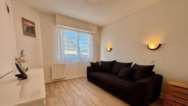 Una sala de estar con un sofá marrón oscuro, una mesa de centro blanca y una mesita de noche de madera. Hay una ventana con cortinas translúcidas, un radiador y una lámpara de pared.