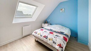 Un dormitorio con techo inclinado, tragaluz, cama con edredón estampado de camiones coloridos, mesita de noche con lámpara y una pequeña decoración en la pared.