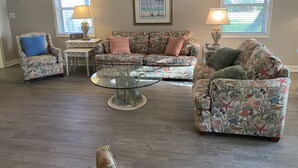 Ein Wohnzimmer mit einem blumengemusterten Sofa, einem Glastisch und einem Sessel mit blauem Kissen. An der Wand hängt ein gerahmtes Bild und auf einem Beistelltisch steht eine Lampe.
