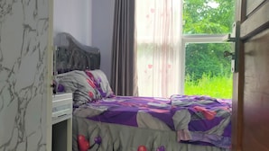 A bedroom with a bed, a nightstand, a window with curtains, and a view of greenery outside. - Azzima villa  (Batu)