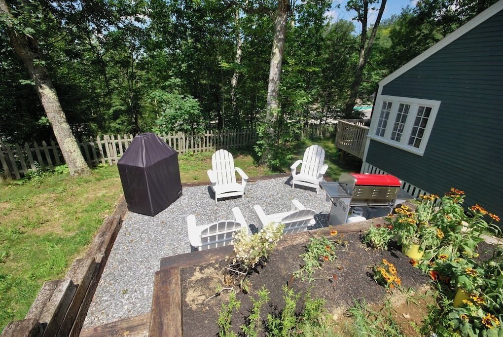 A backyard with a barbecue area, white Adirondack chairs, and a wooden deck.