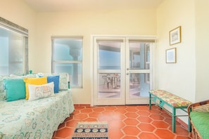 Una habitación con un sofá estampado, un banco y una puerta que da a un espacio exterior con una mesa y sillas.