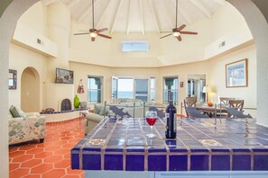 Relax in this spacious living area with a high ceiling, featuring a comfortable seating area and dining table for two, plus a large coffee table and ocean views.