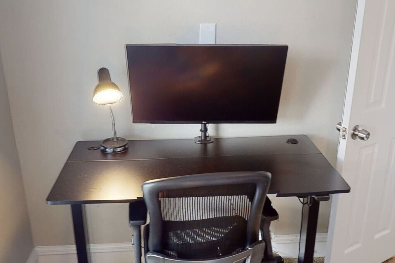 A small office space with a desk, a computer monitor, a chair, and a desk lamp.