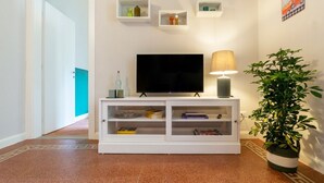Una sala de estar moderna con un televisor de pantalla plana, un mueble blanco para equipos electrónicos, una lámpara de pie y una planta en maceta.