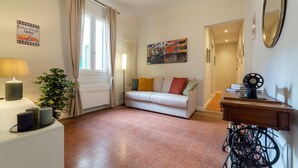 Una sala de estar con un sofá blanco, una lámpara, una planta en maceta, una máquina de coser y un cuadro en la pared.