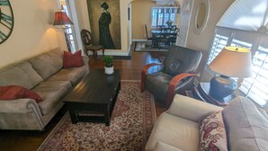 Una sala de estar con sofá, sillón, mesa de centro y alfombra. Hay un cuadro en la pared, una zona de comedor al fondo y una lámpara sobre una mesita auxiliar.