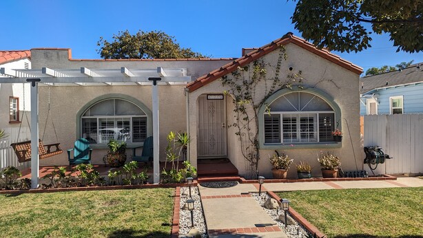 Una casa con un porche cubierto, una puerta principal y un pequeño jardín con plantas en macetas y una silla colgante.