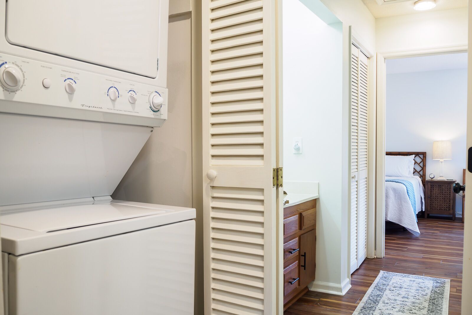 A laundry area with a front-loading washer and dryer, a wooden cabinet, and a bedroom with a bed, nightstand, and lamp.
