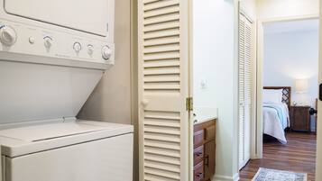 A laundry area with a front-loading washer and dryer, a wooden cabinet, and a bedroom with a bed, nightstand, and lamp.