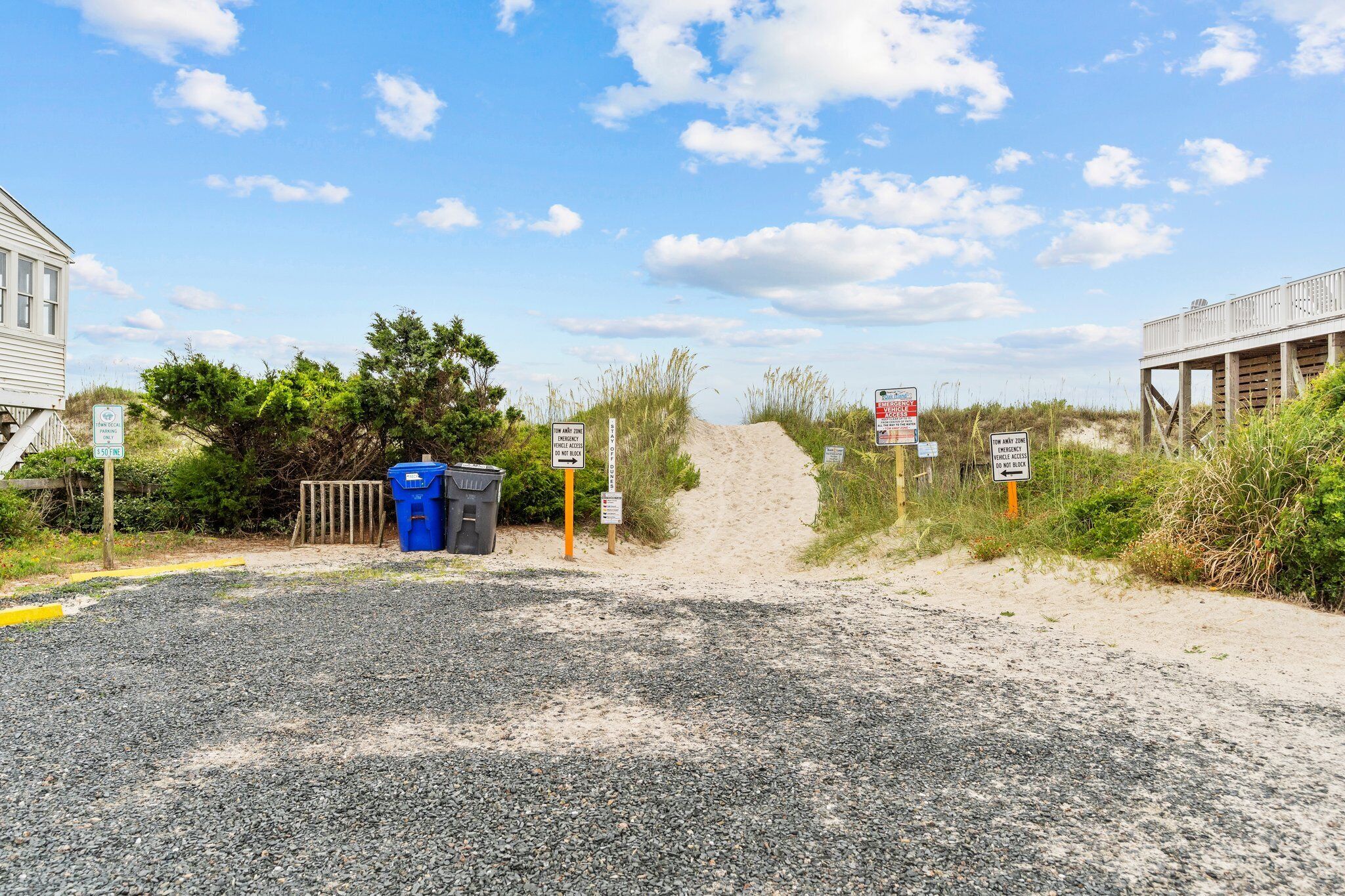 A gravel path leading to a sandy beach area with a wooden structure on stilts, a blue trash bin, and multiple informational signs.