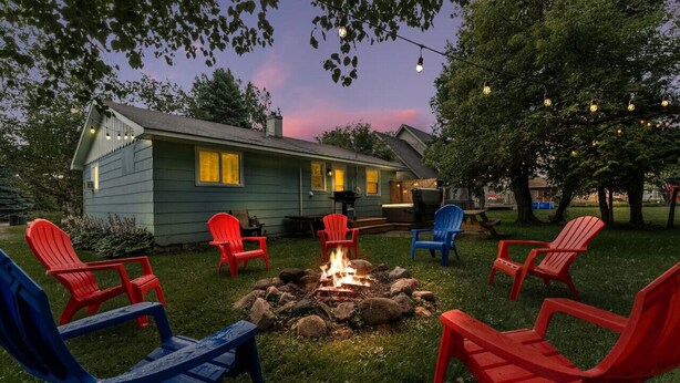 A cozy outdoor seating area with Adirondack chairs around a fire pit, a house in the background, and string lights overhead. - Rock n Roll! DT 2bd home! Walk to beach, bars&more (Port Sanilac)