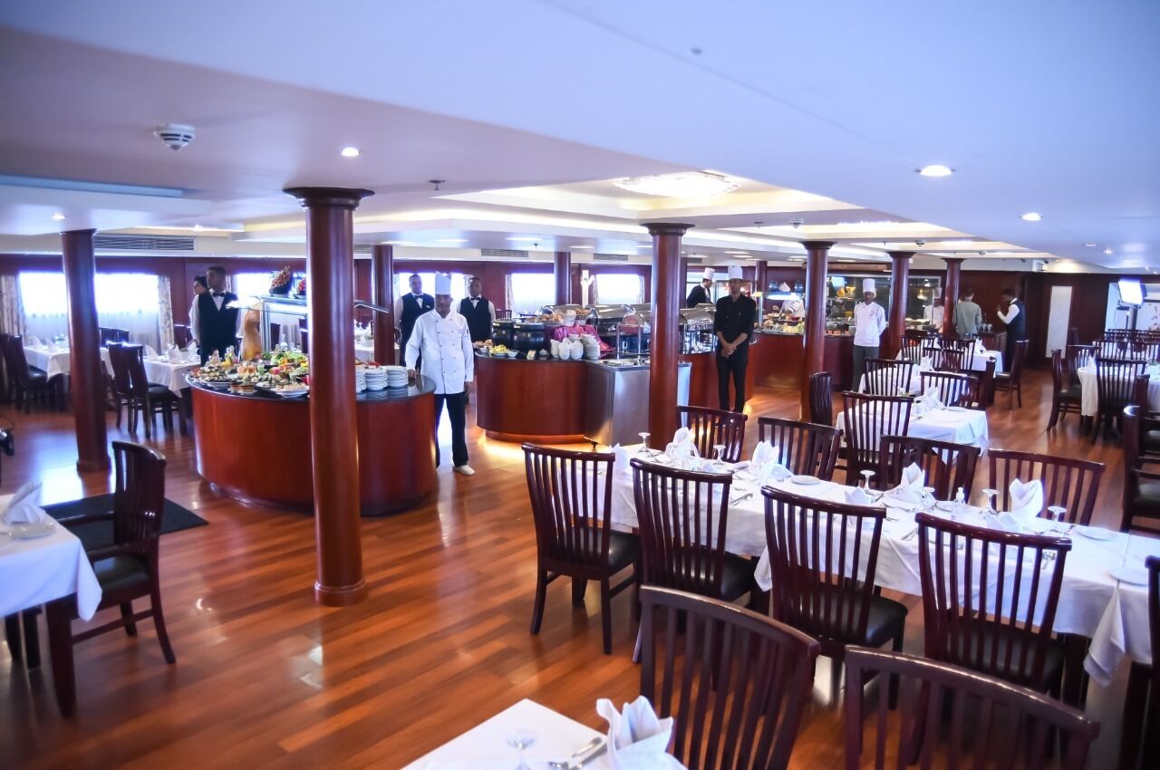 A spacious restaurant with wooden flooring and high ceilings, featuring multiple dining tables and a buffet area.