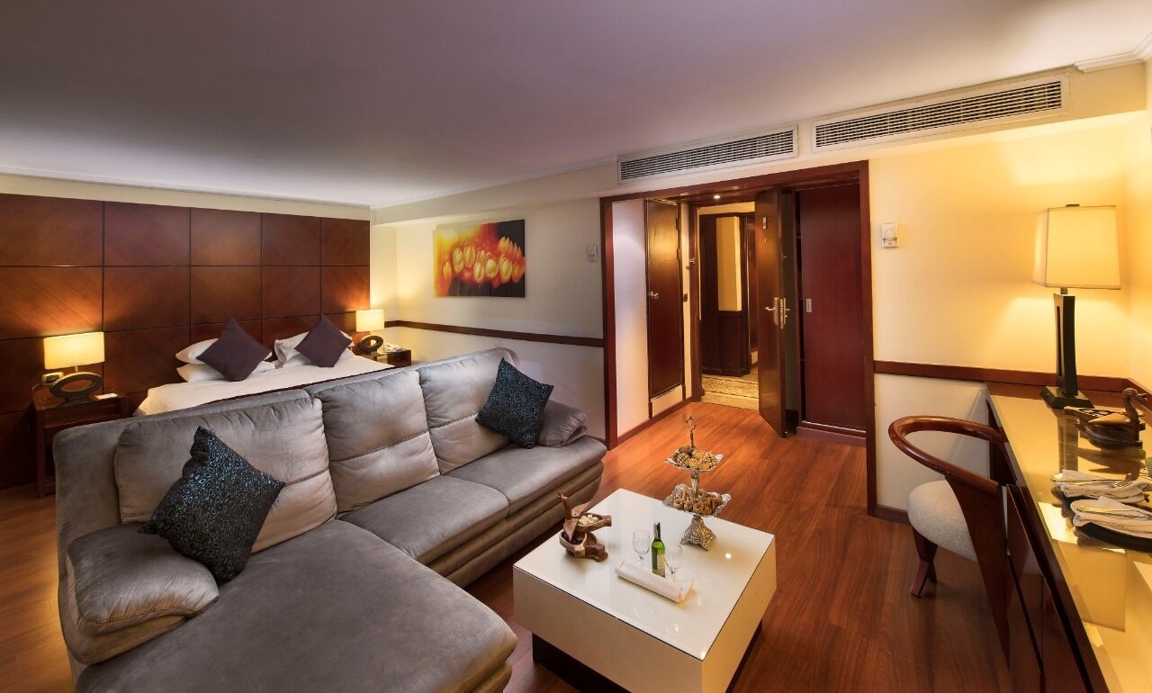 A modern hotel room with a large L-shaped gray sofa, a small white coffee table, and a wooden floor. There is a door leading to a hallway and a room with a lamp and a chair.
