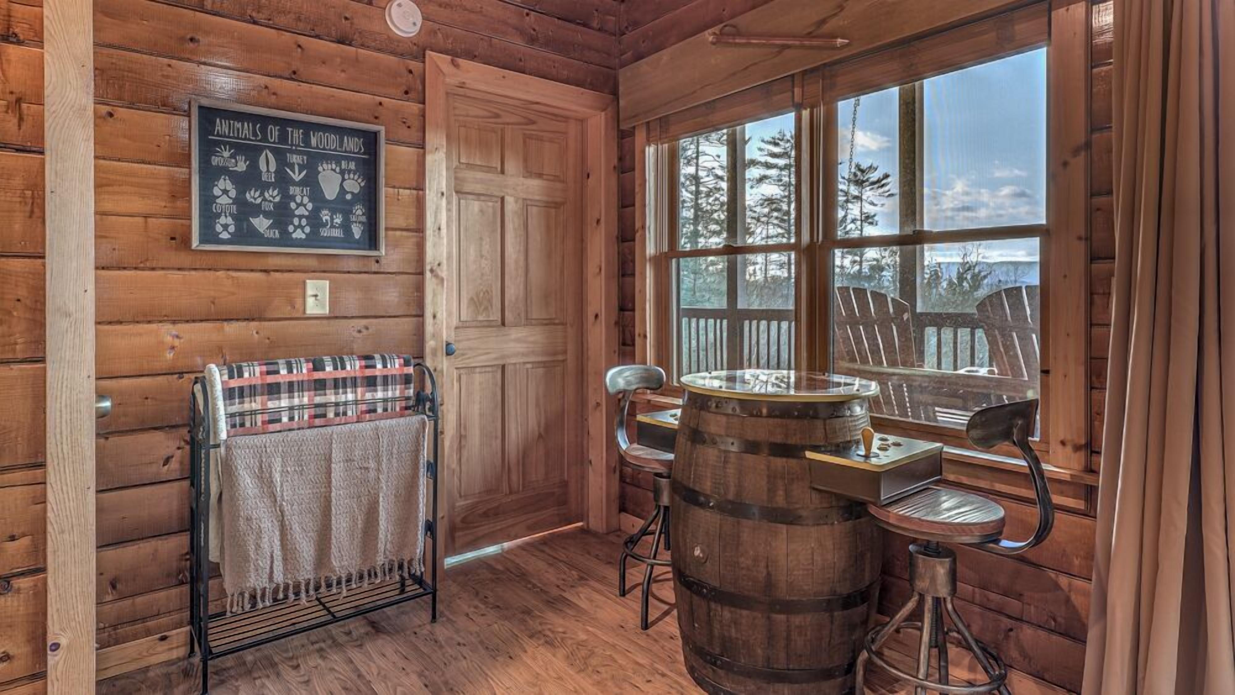 A wooden cabin interior with a wooden wall, a window with a view of trees, a sign listing animals of the woodlands, a plaid blanket on a rack, and a wooden barrel stool.